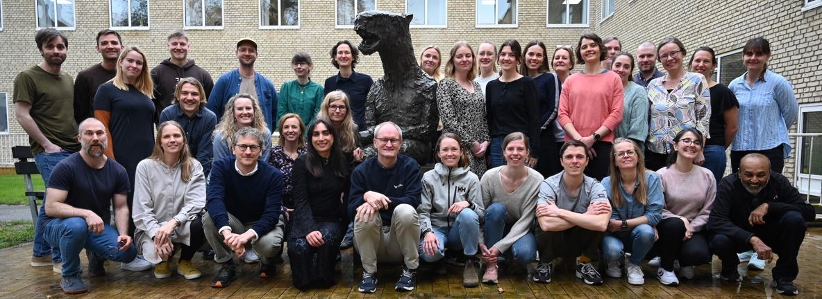 Gruppefoto af medarbejdere ved Forskningsenheden for Almen Praksis foran de karakteristiske gule bygninger ved Institut for Folkesundhed, Aarhus Universitet.